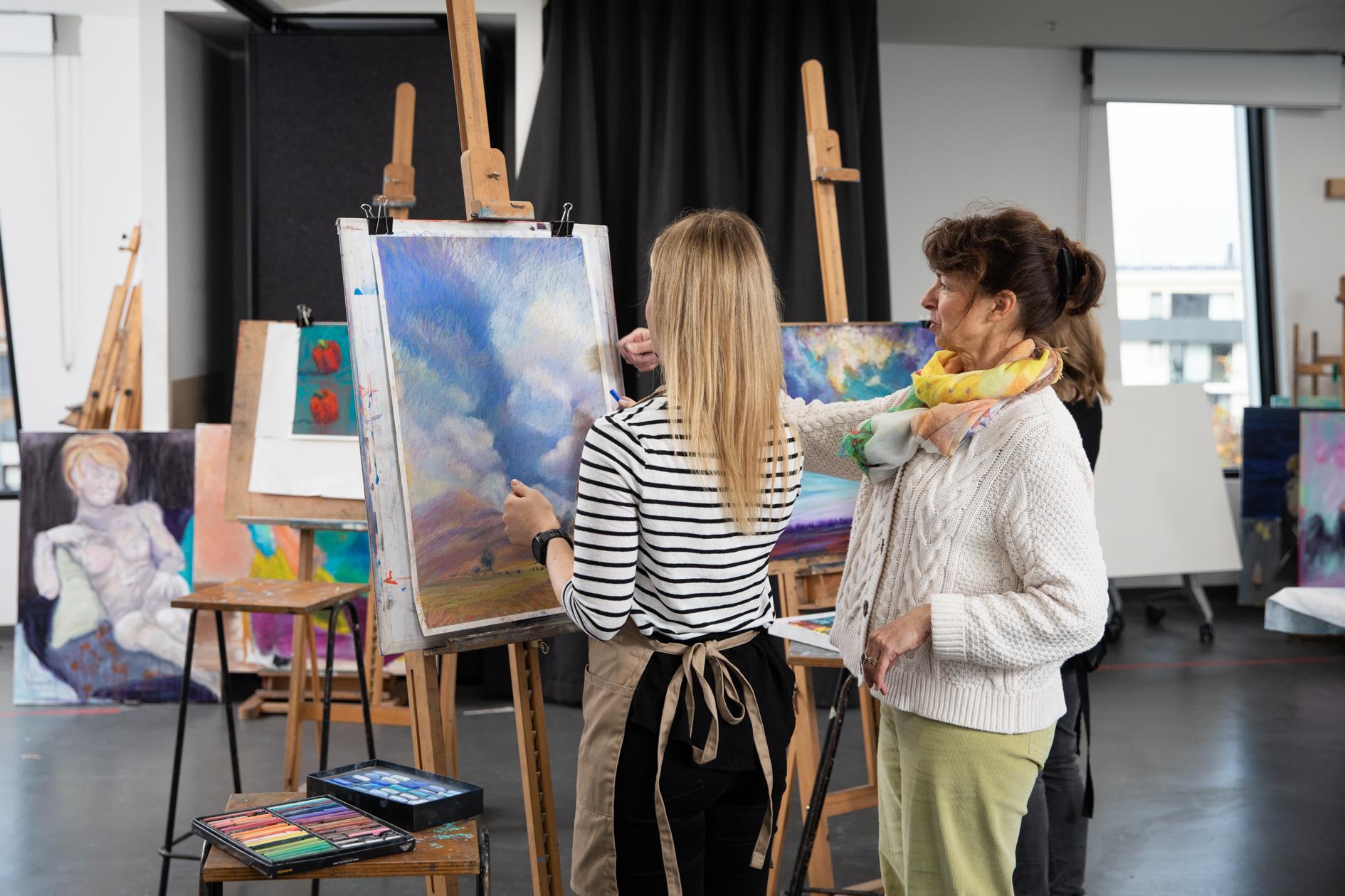 Photo of a student painting a canvas in the centre of frame with a tutor to the right providing instruction.