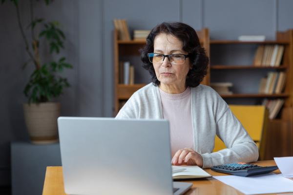 Older woman with dark hair sits at a table using a laptop, there is a calculator and paperwork in front of her.