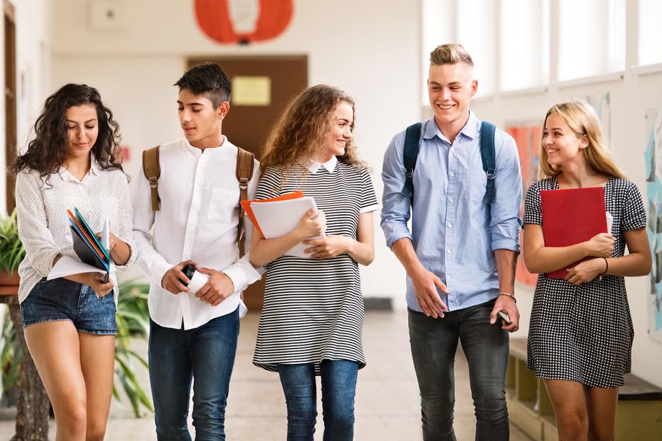 Five young adults carrying books, walking down a wide hallway, smiling and looking at each other