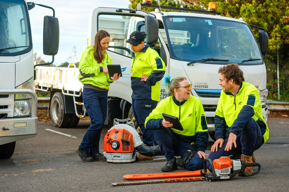 Two women stand at the front of a small truck looking at a mobile device, in front of them a woman and a man are squatting on the ground laughing while looking at a device. Everyone is wearing yellow safety clothing and boots and there are large power tools on the ground near them.