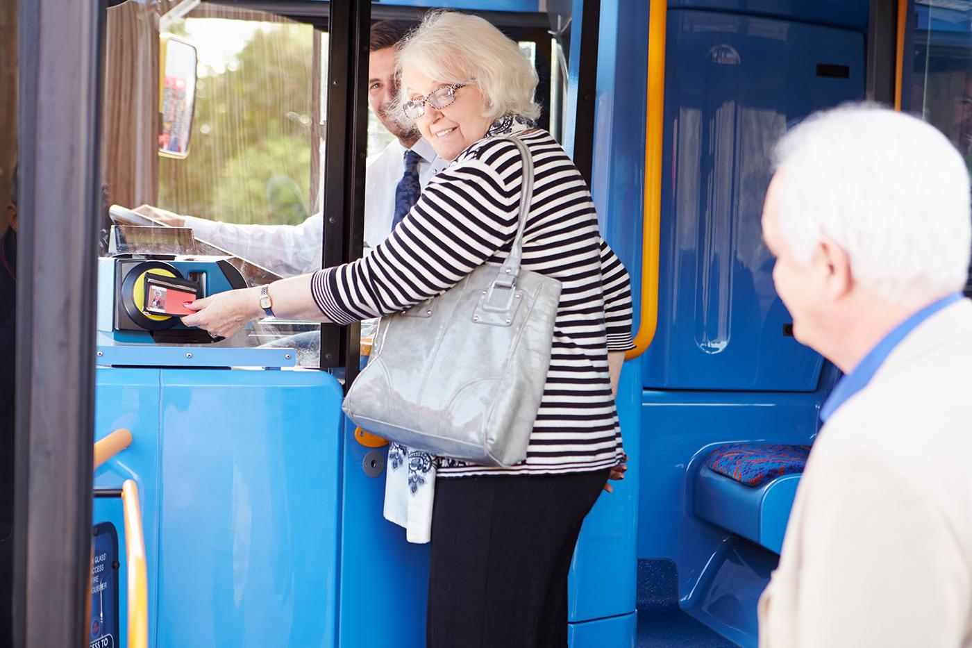 An older woman stands at the entrance of a bus tapping on with her pass, the back of an older man's head is in the foreground.