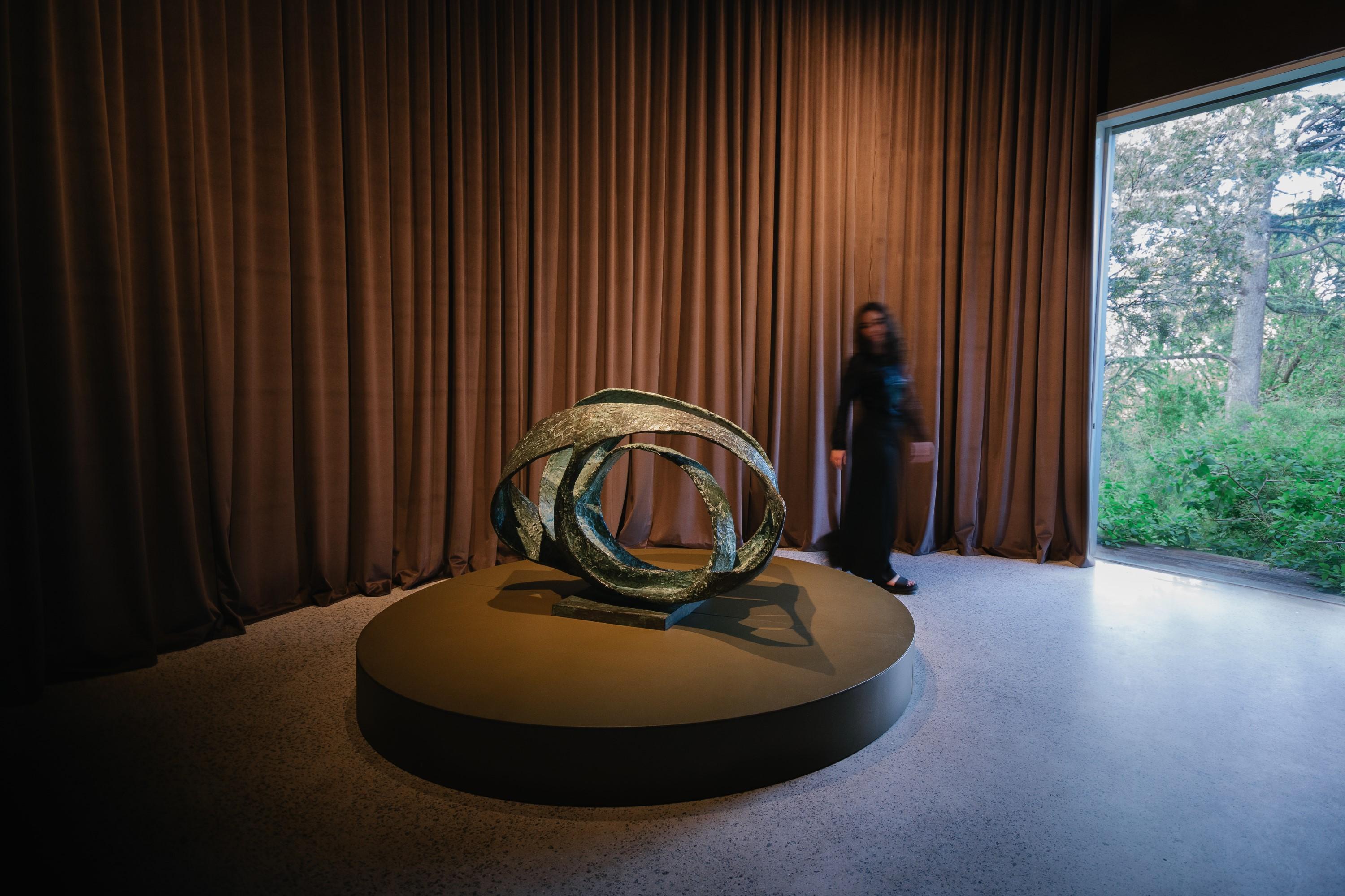 Gallery interior with large window to the right and a dark brown curtain covering the rear wall. A woman dressed in black walks around a a twisted metal sculpture that sits on top of a short circular stand.