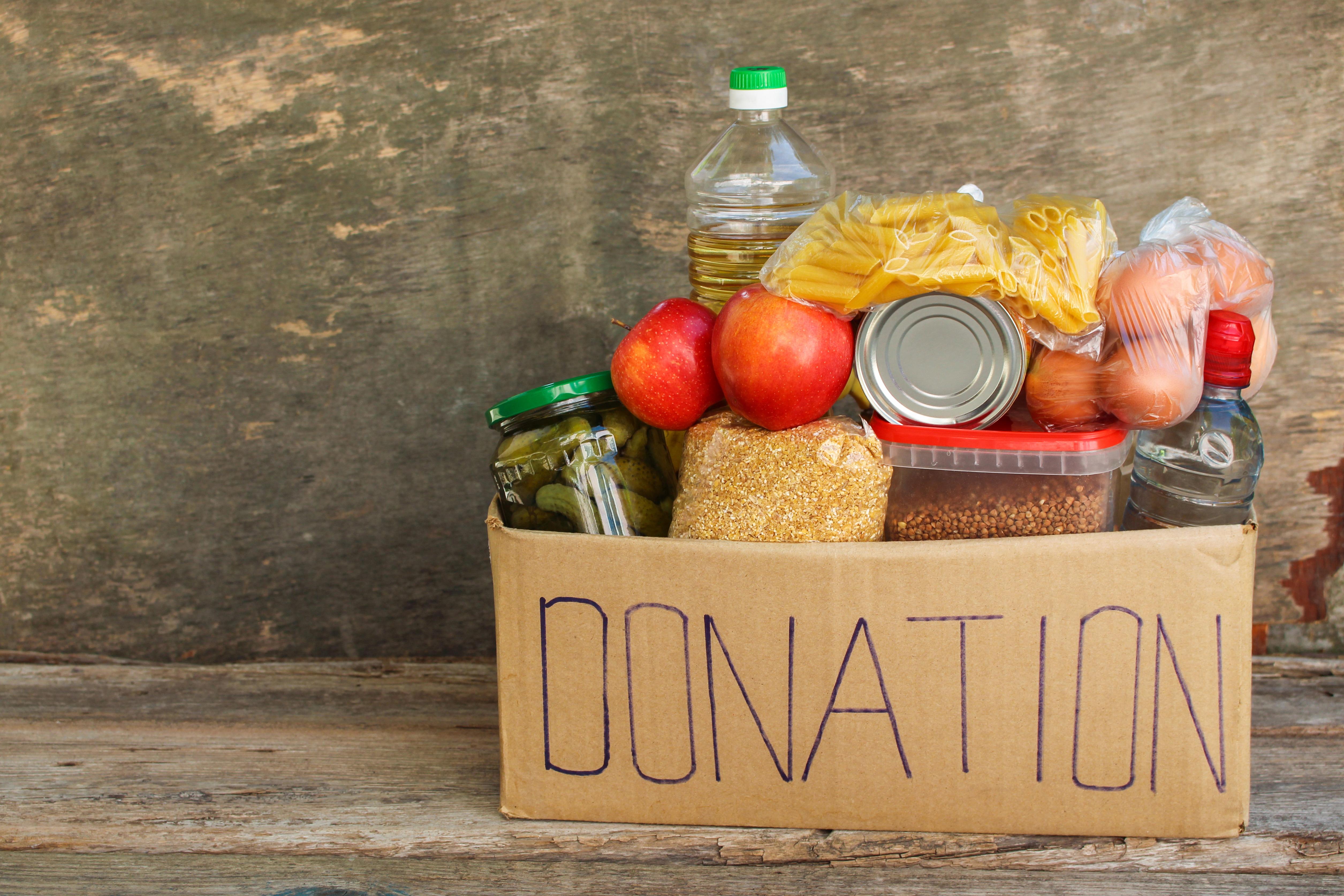 Cardboard box full of food, with the word donation written in blue, sits on a wooden bench..