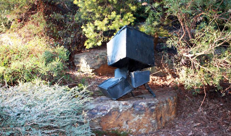 A roughly welded black steel sculpture made up of different sized cubes sits on a rocky step amongst native bushland