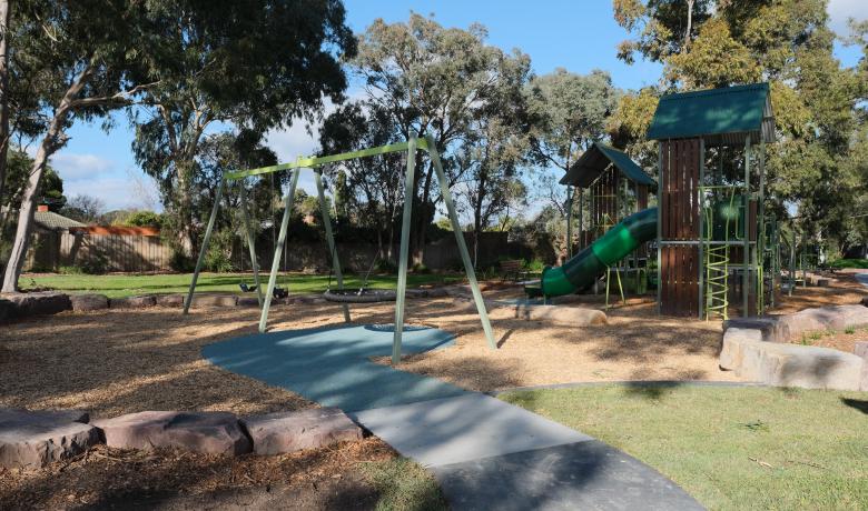 Medium sized playground with swings and a slide sits in a bushy park surrounded by houses.