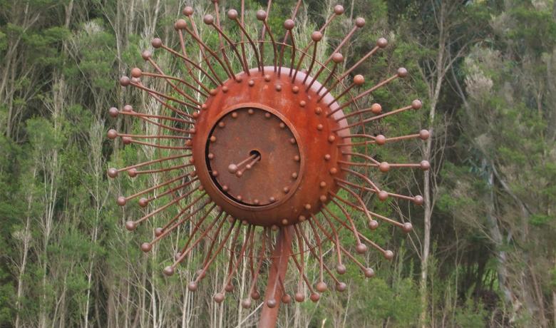 Rust coloured steel sculpture in the shape of a eucalypt blossom sits in a bushy landscape