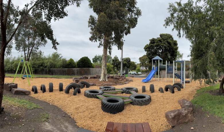 A suburban playground with native trees dotted around, an obstacle course made from tyres, a swing set and a blue slide. Cars and houses can be seen in the distance.