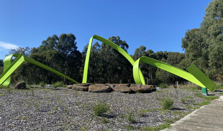 Long twisting lime green steel sculpture sits in a bushy landscape with a concrete footpath off to the right side