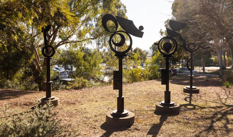 Four tall totemic steel sculptures sit in a clearing amongst bushy native trees, a carpark is in the background.