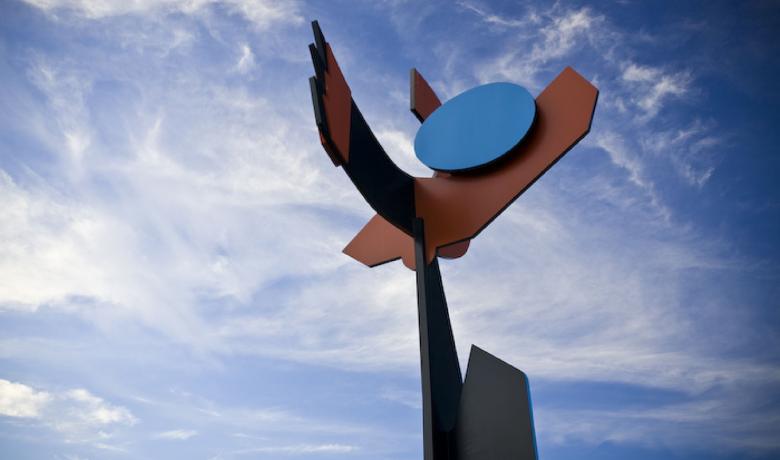 Large steel sculpture comprising bold geometric shapes in black, brown and blue sits against a blue sky.