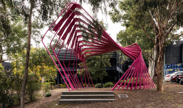 A large pink sculpture made of many bent steel poles sits in a bushy garden in front of a modern office building